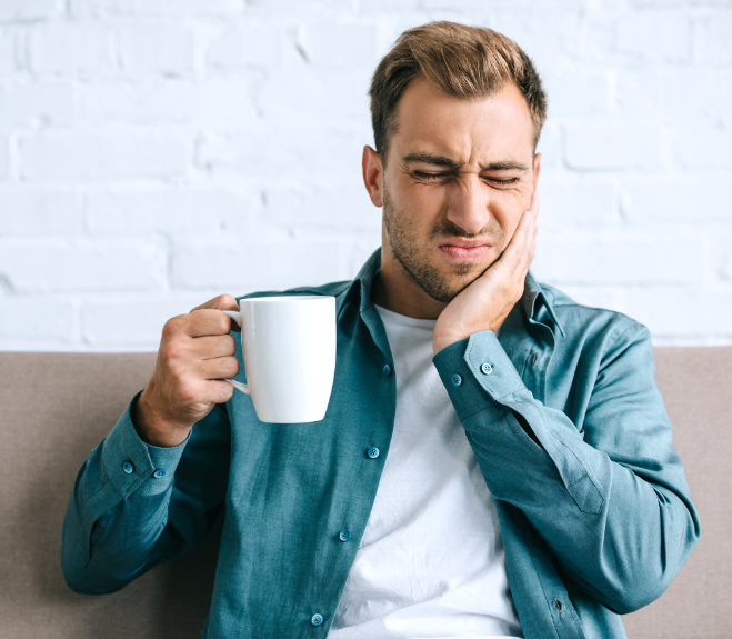 man in turquoise shirt holding a white coffee mug while clasping his jaw grimaces in pain