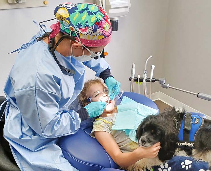 Dr. Bass performs a routine dental procedure on a young girl while Fender the therapy dog comforts the patient.