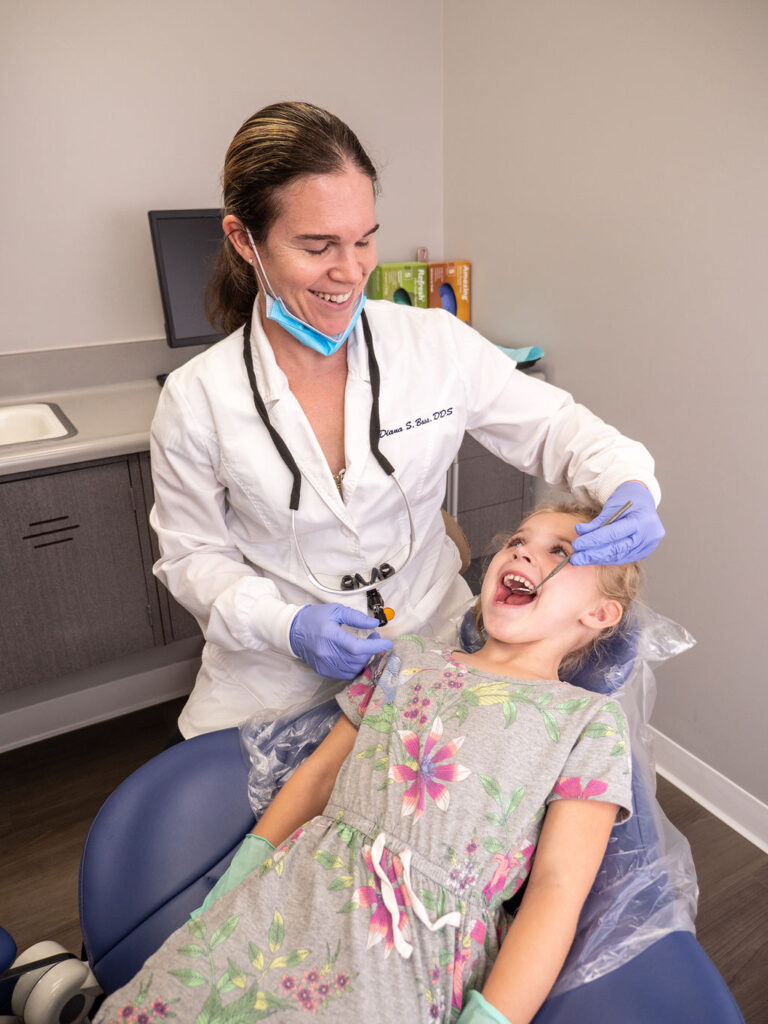 A smiling dentist in a white coat and gloves examines a happy young girl’s teeth in a modern dental office. The child, dressed in a floral dress, reclines in the dental chair while the dentist gently checks her mouth. This image represents general and family dentistry, showcasing a comfortable and welcoming environment for patients of all ages.