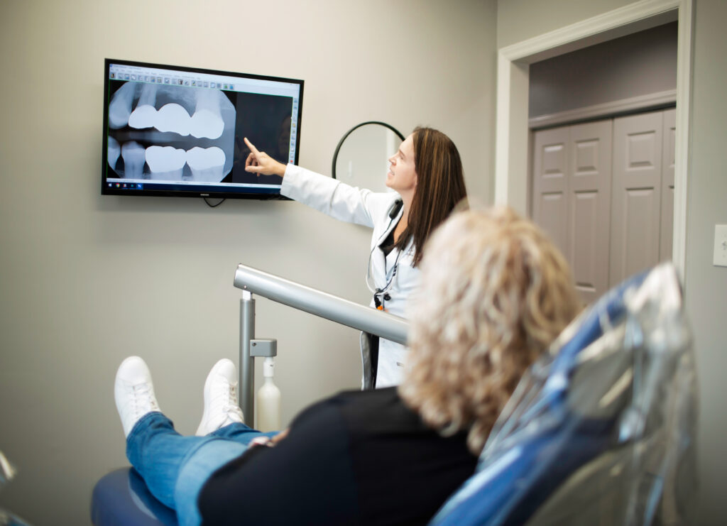 A cosmetic dentist discusses a patient's cosmetic restoration treatment plan, pointing at a digital X-ray displayed on a monitor. The patient, seated in a dental chair, attentively listens while the dentist explains potential procedures for enhancing their smile. The modern office setting highlights advanced dental technology and personalized care.