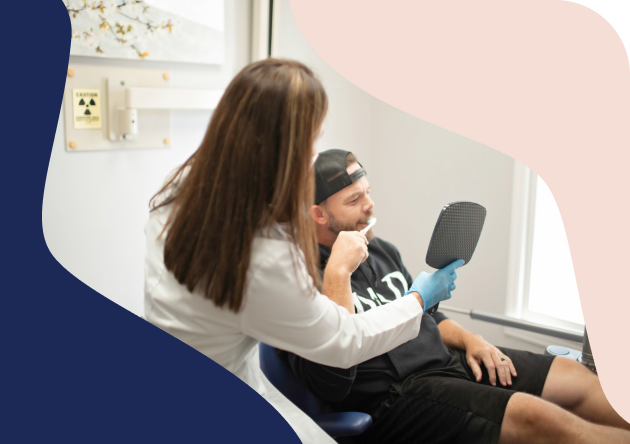 A dentist consults with an adult male patient as he examines his smile in a handheld mirror, illustrating personalized general and family dentistry services with a focus on adult dental care.