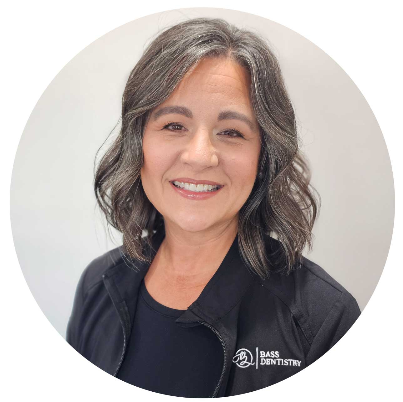 Smiling dental hygienist Nicole with wavy gray-streaked hair wearing a black Bass Dentistry jacket in a professional team headshot